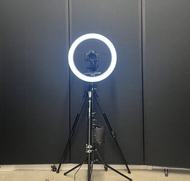 A camera tripod and ring light set up in front of a black backdrop.