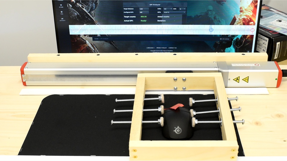 Photo of our CPI testing setup on a wooden desk. A SteelSeries mouse is secured in a wooden clamping frame with metal bolts mounted on a black mousepad. The clamping frame is attached to a linear motion rig with a motor. In the background, a monitor displays the CPI Analyzer tool.