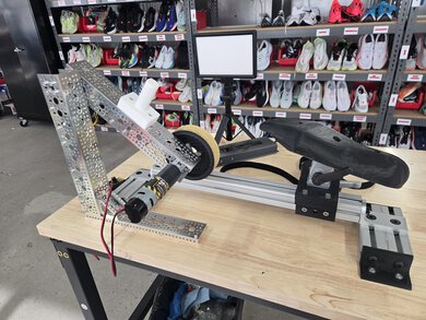 Outsole durability test rig with a motorized sandpaper abrasion wheel, a 3D-printed shoe sole mold on a sliding rail, and an LED light panel, with running shoes on shelves behind.