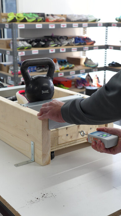 Running shoe outsole traction test using a wooden sled weighted with a kettlebell, pulled by a hand holding a digital force gauge to measure friction, with running shoes on shelves behind.