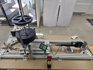 Aluminum shoe traction testing rig with stacked weight plates on a vertical guide rod, a linear actuator and load cell, a touchscreen control panel, an emergency stop button, and a tray of gravel on a wooden workbench.