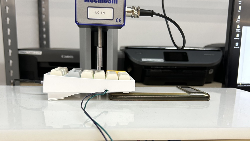 A force testing setup showing a mechanical switch mounted in a modified macropad beneath a vertical force testing probe. The probe is about to press downwards on a keycap to measure actuation force, with wires extending from the macropad for data collection.