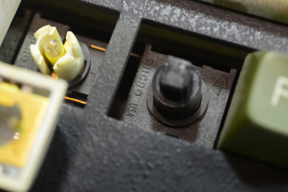 Close-up view of a vintage Hall effect keyboard switch with keycaps removed, showing the exposed switch housing marked “Micro USA.” The switch on the left features a distinctive, center-mounted LED.