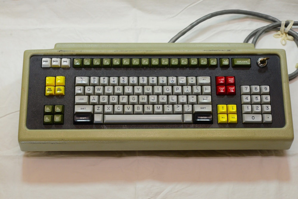 A vintage terminal keyboard from the 1960s or 1970s featuring Hall effect switches. It has an olive-colored case, a built-in numeric keypad, and numerous distinctive function and command keys not found on modern keyboards.
