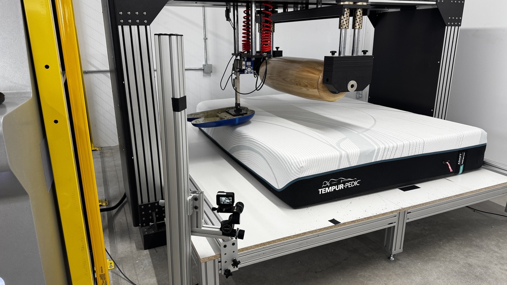 The Tempur-Pedic TEMPUR-Adapt in a mattress testing machine with a large wooden roller and a blue naturalistic loading pad in place to test the mattress's firmness.