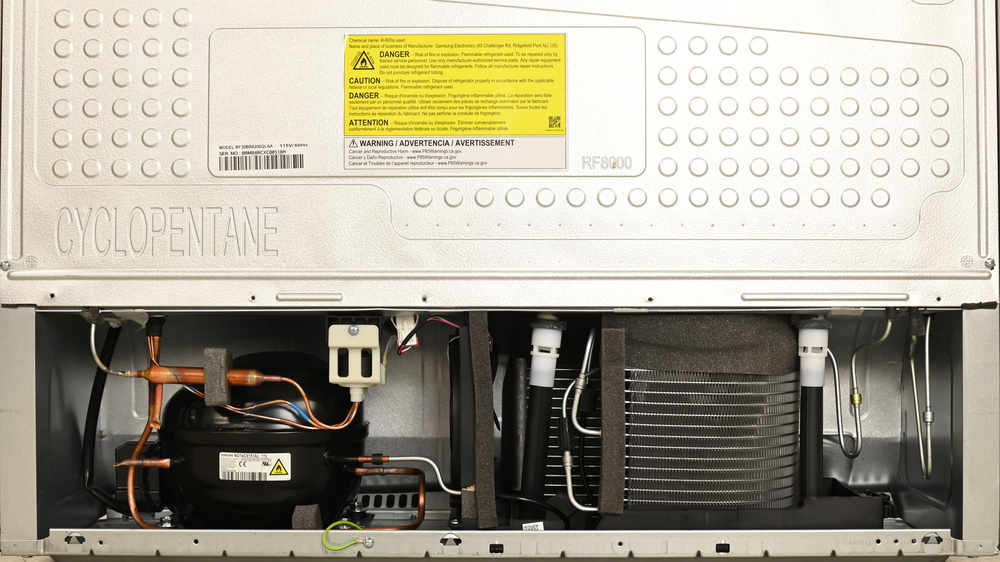 On the right there are two large diameter black tubes coming from the interior above, leading to a shallow, wide black pan below. A large condenser with many fins sits inside the pan. The compressor is on the left and there's a slim horizontally-oriented fan in the center, with its airflow directed left to right.