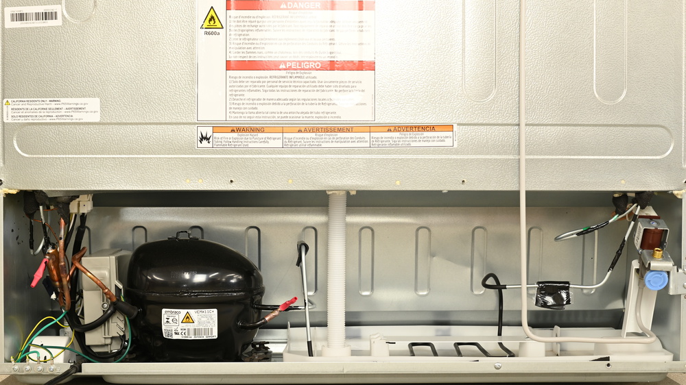 The machine compartment at the bottom rear of the KitchenAid KRQC506MPS, showing a fan on the left cooling an oval black compressor just to the left of center. The silver condenser tube leads from the compressor and is bent into several loops sitting above the floor of a white plastic defrost meltwater tray. On the right is a brass threaded connection for a water line, with a gray rigid plastic tube leading upward.