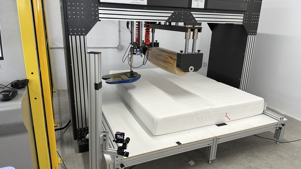 The Sleep On Latex Pure Green Organic in a mattress testing machine with a large wooden roller and a blue naturalistic loading pad in place to test the mattress's firmness.