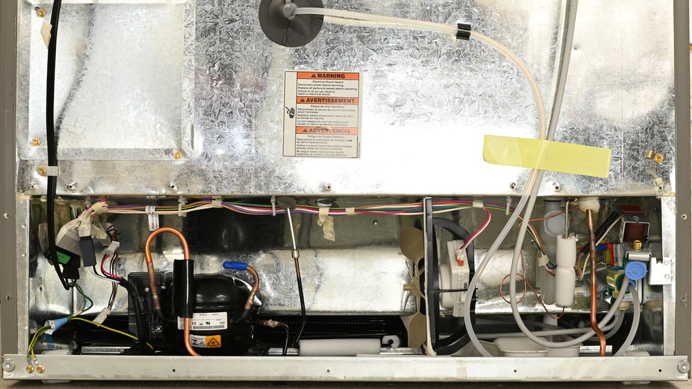 This is a silver galvanized steel machine compartment, with an oval black compressor on the left and a condenser folded underneath the compartment, behind the compressor. The condenser is quite long, extending from the left to the center, and is folded into two loops. There's a beige plastic fan to the right of center, oriented so air blows horizontally across the compartment. There's a threaded water connection on the right, with a clear tube leading to the center back of the fridge section and entering through a sealed opening. Another gray water tube leads up and to the right.