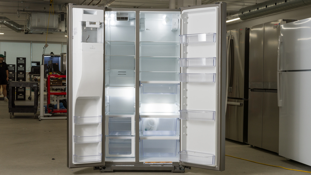 The interior of a side-by-side refrigerator. On the left is a narrow freezer section, with an ice chute covering the top of the door and extending down more than halfway, with three small door bins underneath. There are five shelves in the freezer with two vertically-stacked pull-out drawers on the bottom. The fridge section on the right is a little wider than half the width of the fridge cabinet and consists of three glass shelves, a meat or deli drawer underneath a fourth shelf, and a fifth shelf on the bottom with two crisper drawers vertically-stacked underneath. There are five door bins in the door on the right.