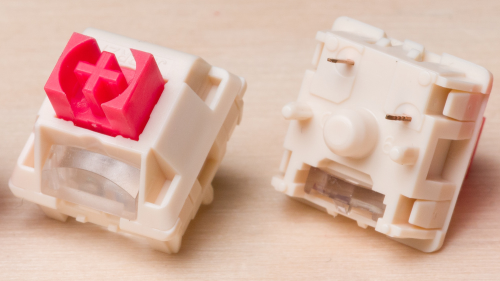 Two mechanical keyboard switches with white housings and red cross-shaped stems shown on a wooden surface, one facing top side and the other bottom side. Both switches feature a clear light guide window built into the housing for improved illumination.