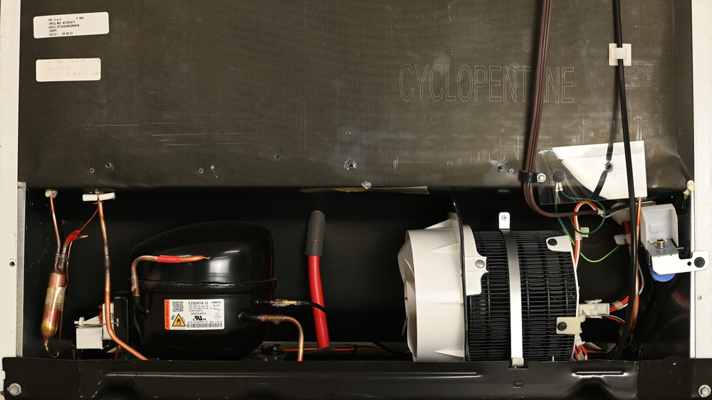 Inside this black machine compartment, from left to right, a copper bulb for the filter dryer, a black oval compressor, a red tube coming from the interior of the fridge above, then a black rolled tubular condenser grid. There's a white fan bolted to the left of the condenser. On the right is a threaded water connection, pointing upwards.