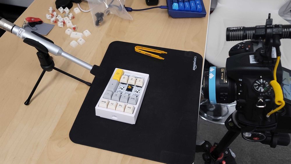 A desk setup showing our Factory Smoothness setup. A small macropad with several keycaps removed sits on a desk mat, with a condenser microphone on a tripod positioned close to it and a DSLR camera mounted on a tripod recording from the side. Nearby are loose keycaps, switches, and a yellow switch puller.
