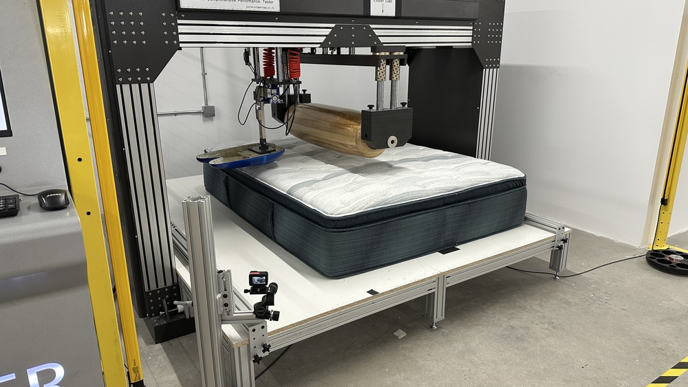 The Beautyrest Harmony Lux in a mattress testing machine with a large wooden roller and a blue naturalistic loading pad in place to test the mattress's firmness.