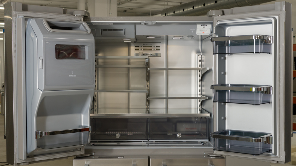 The interior of the refrigeration section of a 5-door refrigerator. The left door consists of an ice chute with a shallow bin underneath. There's an ice maker behind a gray flap at the top of the fridge interior, followed by four half-width shelves, and a full-width fifth shelf with two crisper drawers mounted underneath. There are three door bins in the door on the right.