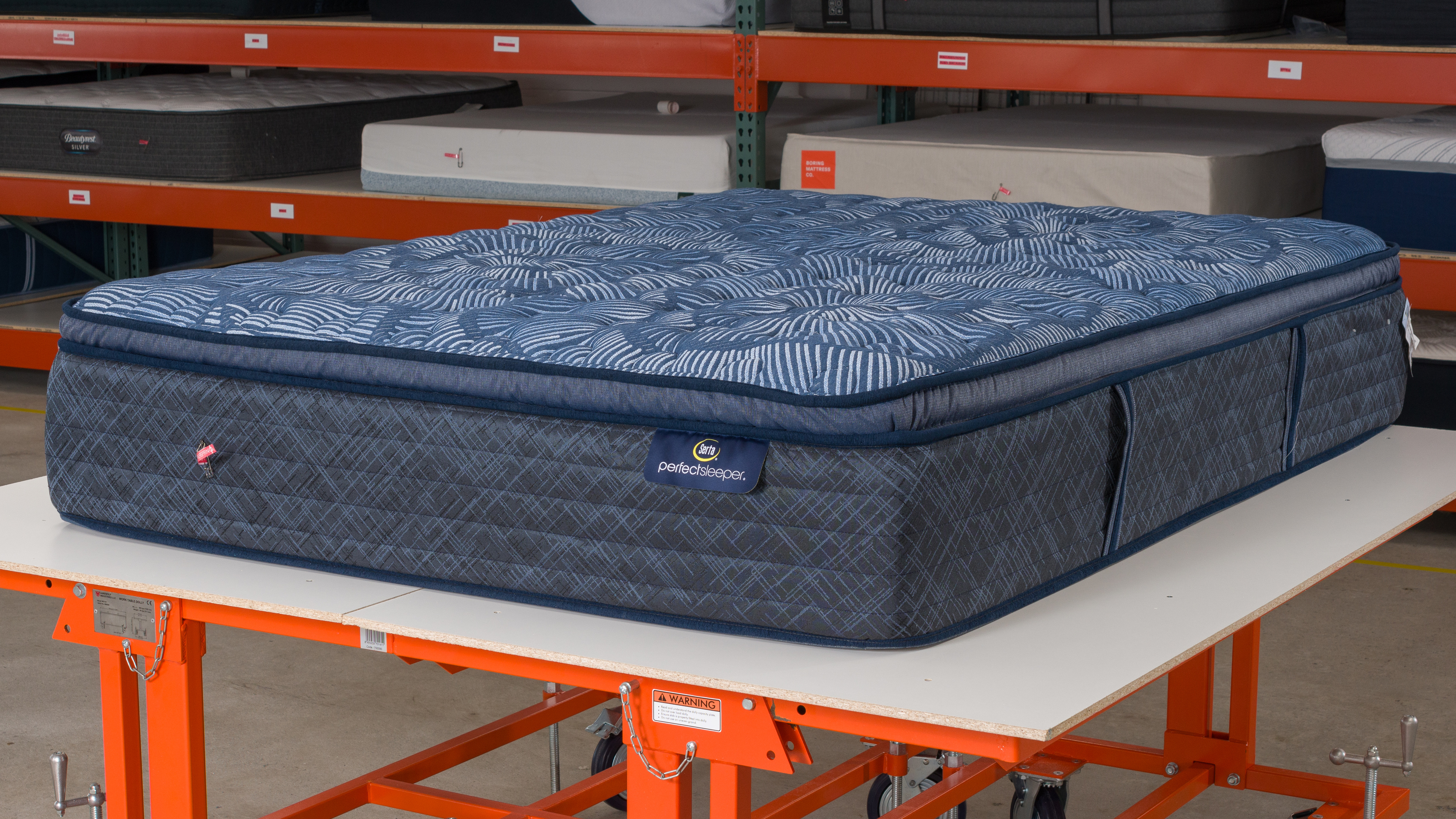 The Serta Perfect Innerspring on a platform in a warehouse-like environment. In the background, there are many other mattresses, stacked on shelves.