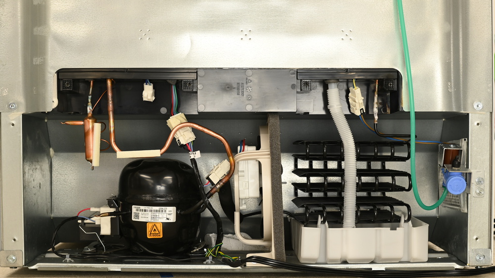 There's a deep plastic tray on the right, with a flexible white plastic tube leading into it from above. The black condenser consists of tubes folded back on themselves several times with fins running in between, sitting on clips just above the tray. The compressor is on the left, with a horizontally-oriented fan in the center of the compartment. The fan's airflow is directed left to right.