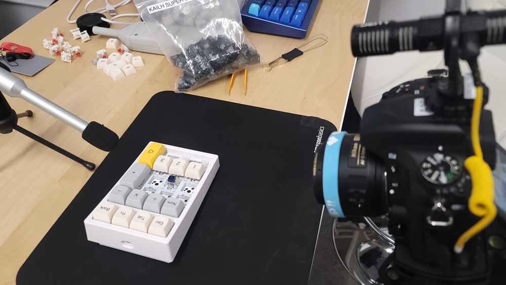An overhead shot showing a white macropad with several switches and keycaps removed. A microphone and a camera on a tripod are positioned nearby.
