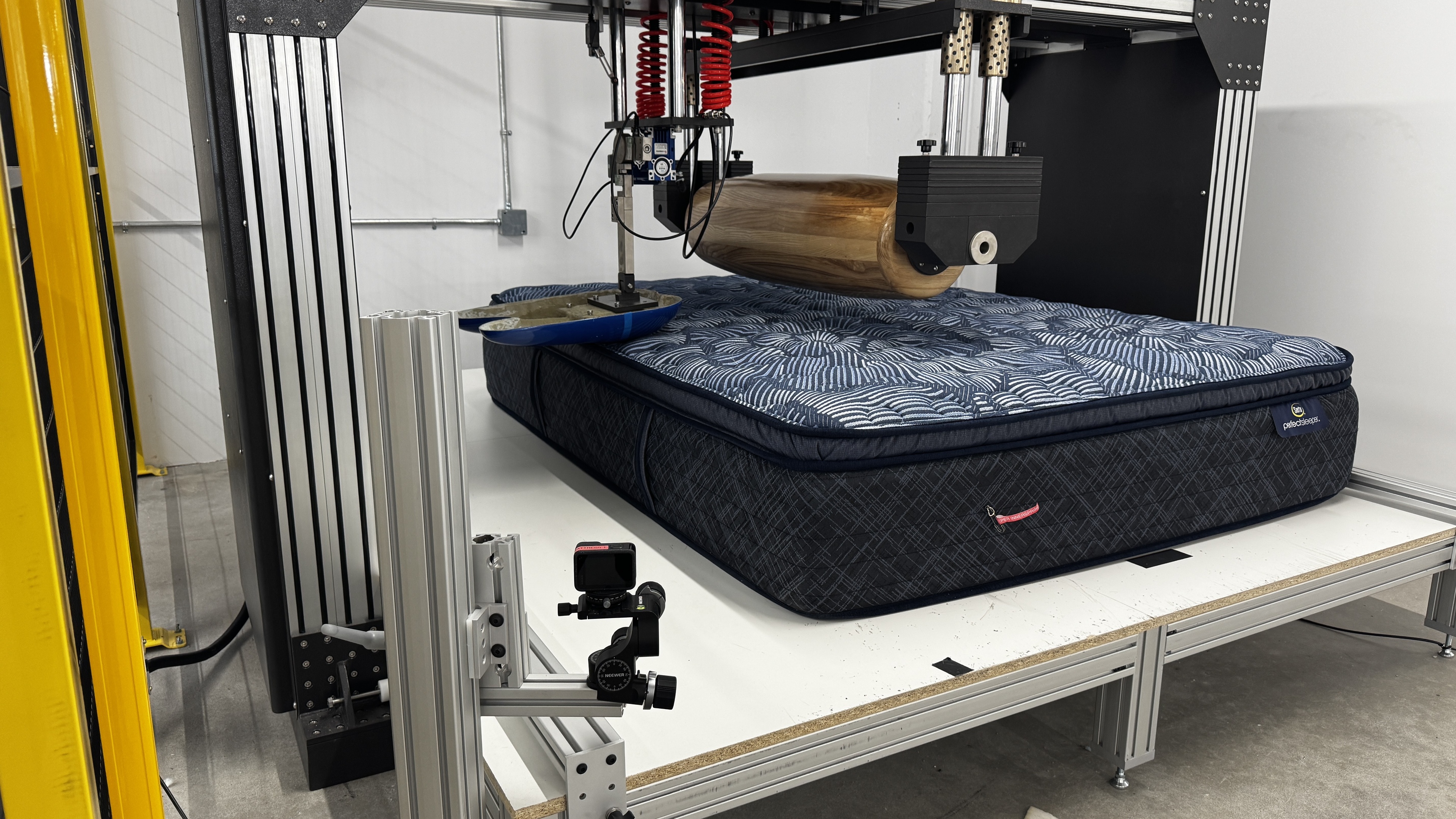 The Serta Perfect Sleeper Innerspring in the mattress firmness testing machine. There's a blue loading pad and large wooden roller above the mattress.