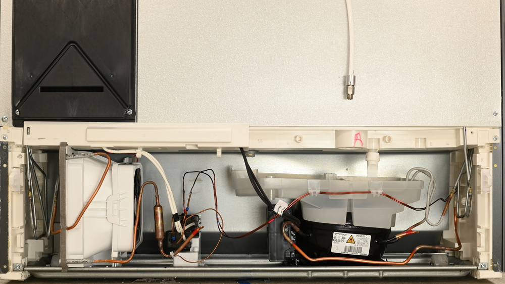 A translucent tray sits on top of a black compressor on the right, with a spout above the tray. Gray waterproofed condenser lines bend into the tray from the right. On the left is a black fan in a white intake shroud.