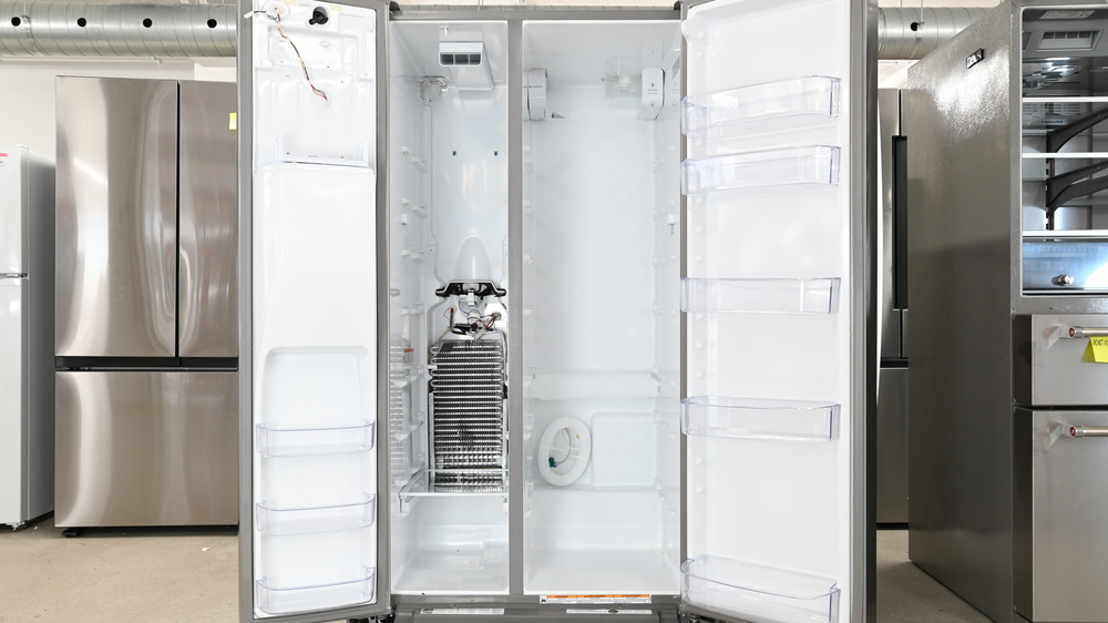 The internal components of a side-by-side refrigerator. All shelves and panels have been removed, exposing the silver evaporator on the left in the freezer section. It's vertically oriented, tall, and narrow. Above it is a fan pointing upward. In the fridge section on the right, there are many loops of white tubing bundled together near the bottom of the fridge at the back.