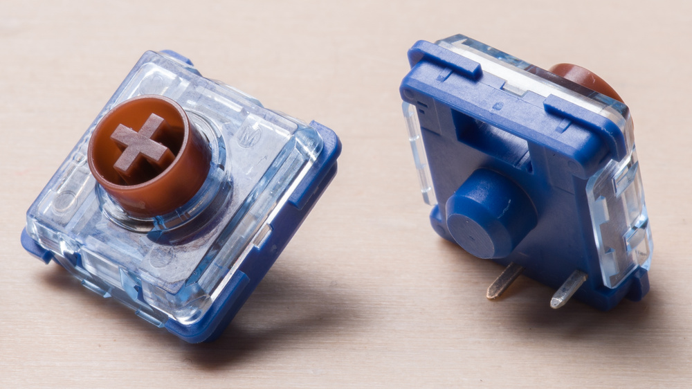 Two low-profile Kailh mechanical switches shown on a wooden surface, one facing top side showing its MX-Style stem shape and the other showing the bottom pins and fairly wide center post.