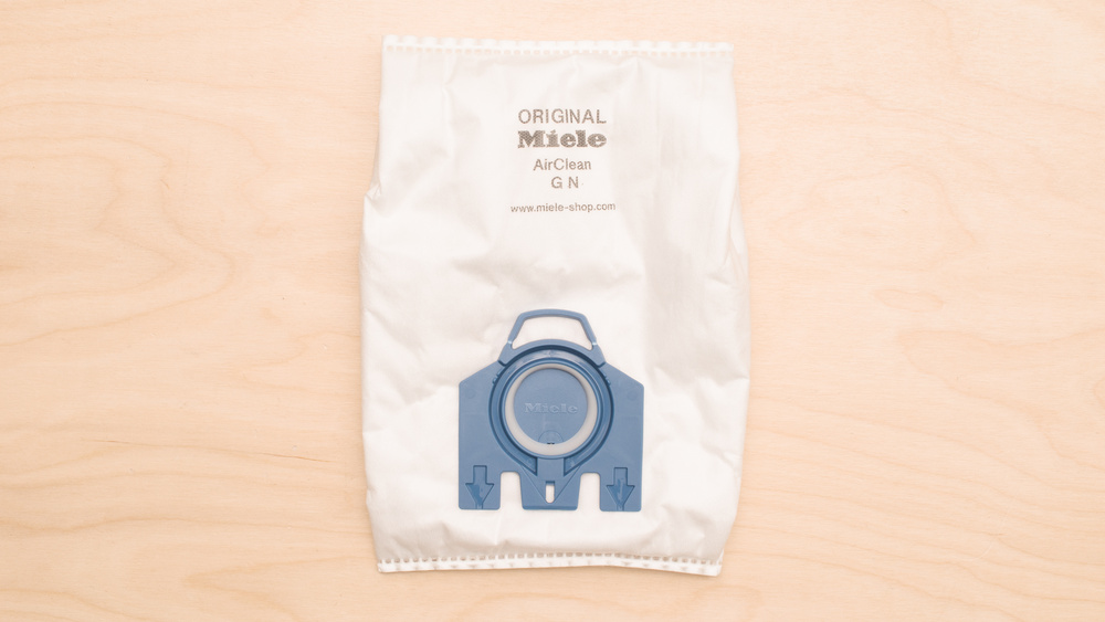 White nonwoven polymer fabric bag with a blue plastic seating plate near the bottom used in the Miele Complete C3 vacuum. There's a blue plastic sealing plug in place over the inlet hole.