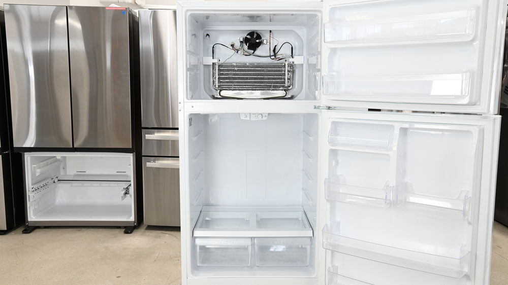 A white top freezer refrigerator with all shelves and panels removed. In the freezer section there's a large silver evaporator with a fan above it, pointing outward.