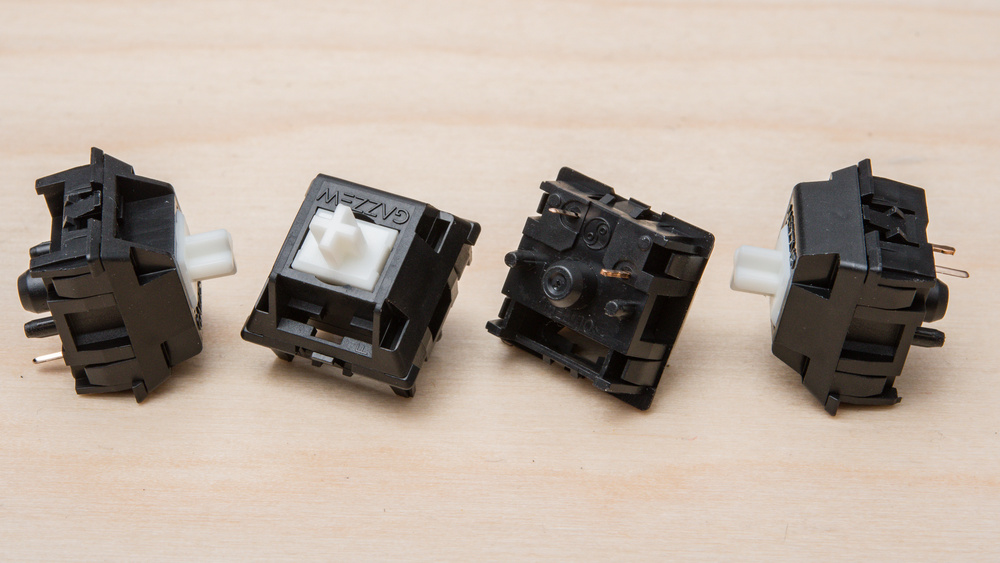 Four of the same mechanical keyboard switches shown on a wooden surface, angled to display different sides. The switches have black housings with white cross-shaped stems and visible metal pins and plastic stabilizer pins on the underside.