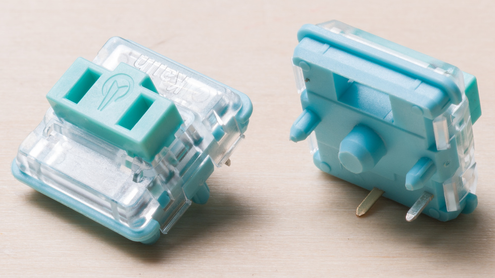  Two low-profile Kailh mechanical switches shown on a wooden surface, one facing top side showing its Choc V1 stem shape and the other showing the bottom pins and fairly narrow center post.