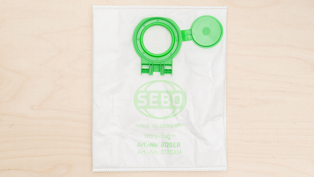 White nonwoven polymer fabric bag with a green plastic seating bracket at the top of the bag used in the SEBO Airbelt D4 vacuum. There's a green flap attached to the seating assembly using a flexible plastic hinge.