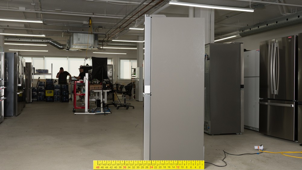 The gray side of a stainless steel-fronted refrigerator with a dimensioned stick positioned to measure depth. The depth measures a little over 27".