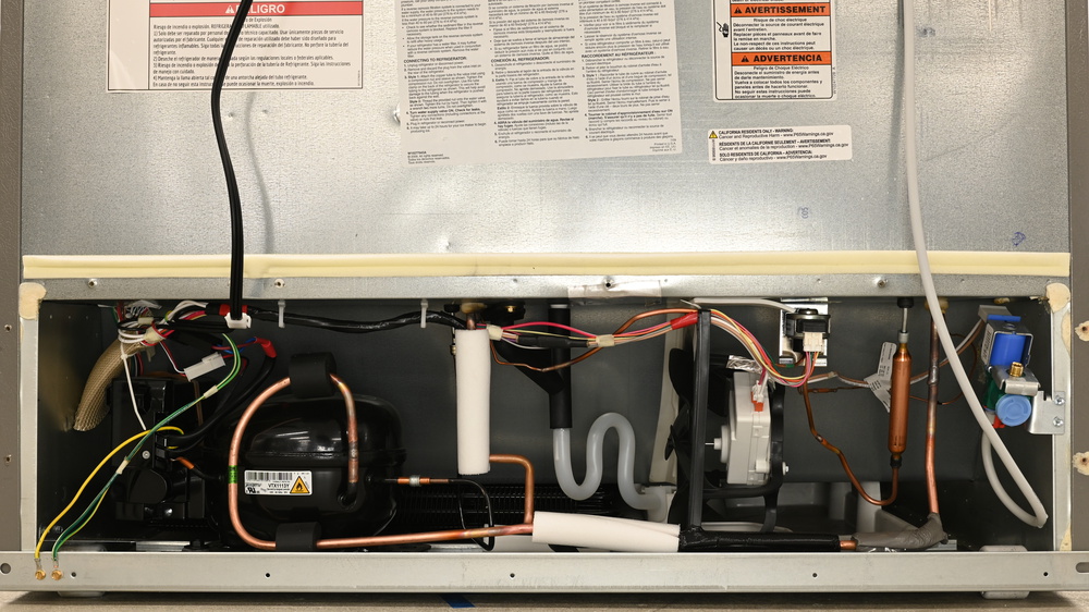 The machine compartment at the bottom rear of the KitchenAid KRMF706ESS, with the oval black compressor on the left. Thick copper tubing exits the compressor. The condenser is folded in two layers and is positioned just behind the compressor, extending underneath the freezer compartment. A black Y-connection connects two defrost drain lines to an S-shaped line that connects to a pan behind a fan on the right. The threaded water connection is on the right, which feeds a flexible water line running upward.