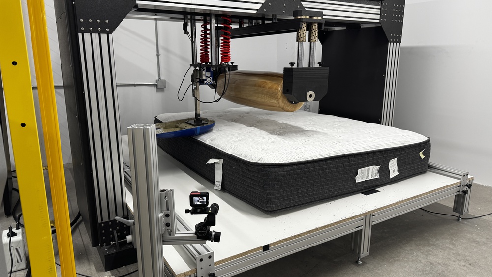 The Beautyrest Silver BRS900 in a mattress testing machine with a large wooden roller and a blue naturalistic loading pad in place to test the mattress's firmness.